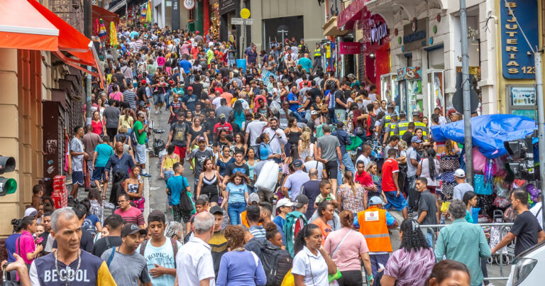 A imagem mostra uma rua bastante movimentada, cheia de pessoas caminhando em ambas as direções, carregando sacolas de compras. As fachadas das lojas têm letreiros coloridos