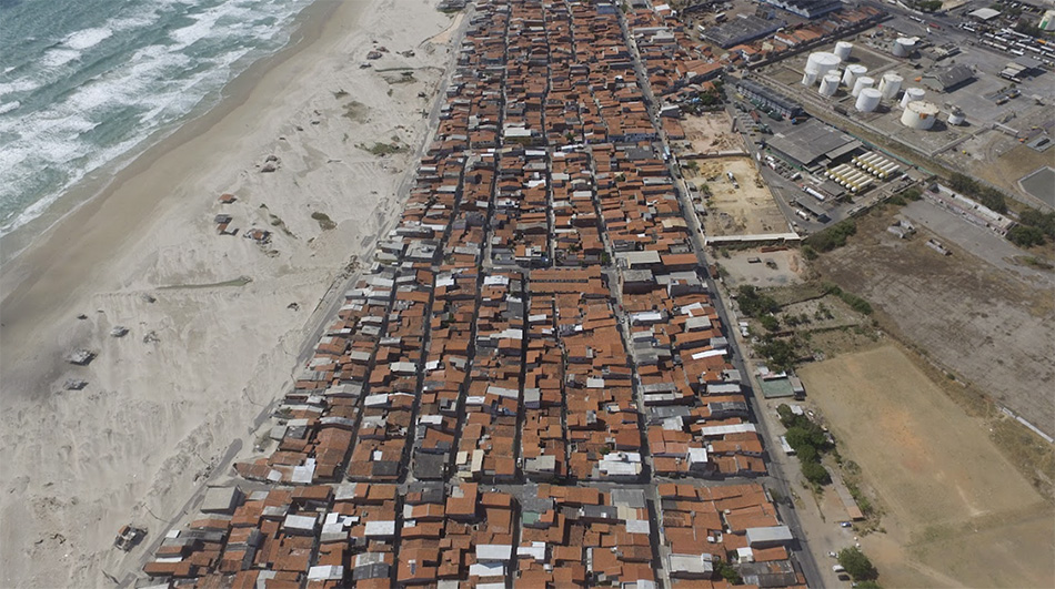 A foto é a vista aérea da comunidade urbana Serviluz, construída na faixa litorânea. À esquerda, vemos o mar com ondas e a faixa de areia. À direta, várias casas dispostas sobre a faixa de areia.