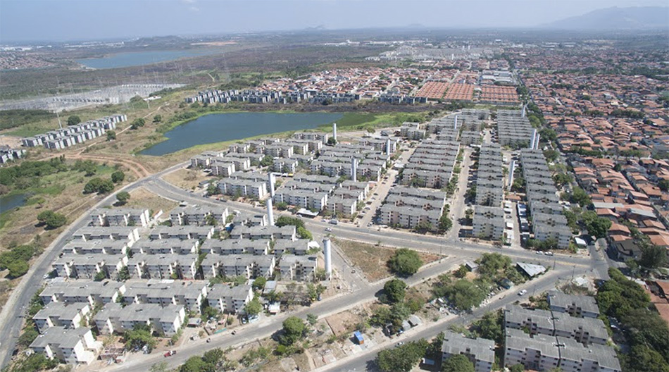 A imagem é uma vista aérea do Conjunto Habitacional Cidade Jardim. Ábaixo e à esquerda. estão vários prédios cinzas; à direta, casas com telhas. Acima, à esquerda, visualizamos um lago.