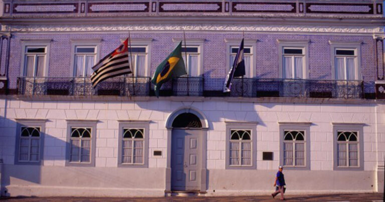 A fachada do Museu Republicano de Itu tem porta e janelas de madeira, paredes revestidas por azulejos e três bandeiras penduradas sobre a porta: a do estado de São Paulo, a do Brasil e a da USP.