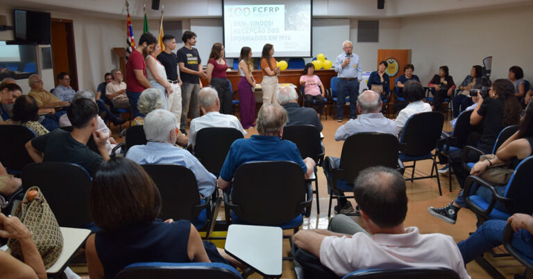 Na imagem aparecem alunos e ex-alunos da Faculdade de Ciências Farmacêuticas, em primeira plano alguns sentados e ao fundo alunos em pé a frente de um telão