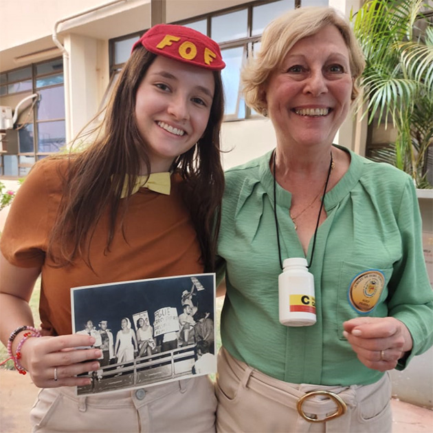 Na imagem aparecem duas mulheres brancas, uma jovem de cabelos pretos e compridos e com uma boina vermelha na cabeça e uma de meia idade loira de cabelos curtos 