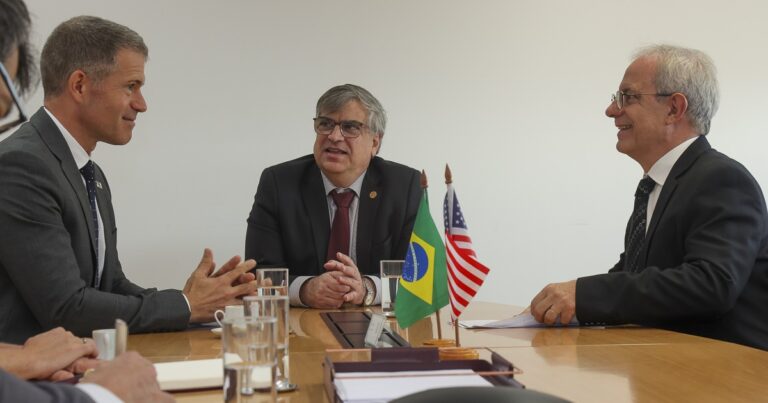 Sentados à uma mesa de madeira, três homens, de terno. Sobre a mesa, papéis, xícaras e pequenas bandeiras do Brasil e dos Estados Unidos