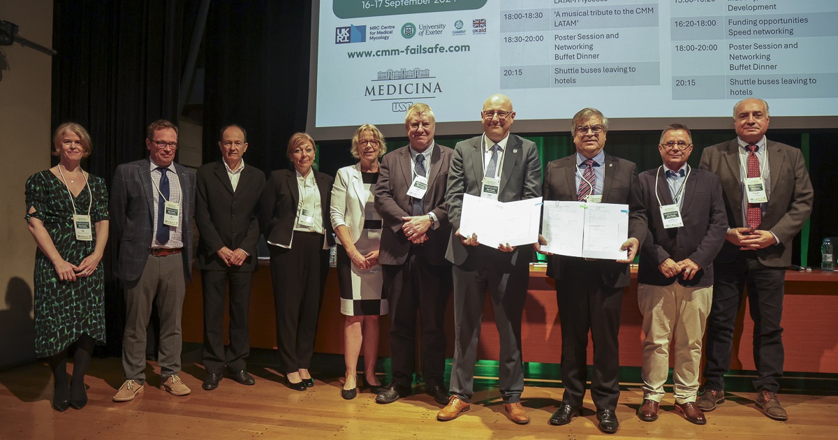 Em um palco de madeira, 10 pessoas estão em pé, posando para a foto. Duas delas seguram uma pasta aberta, mostrando um documento, que não é legível na foto. Ao fundo, uma mesa e um telão