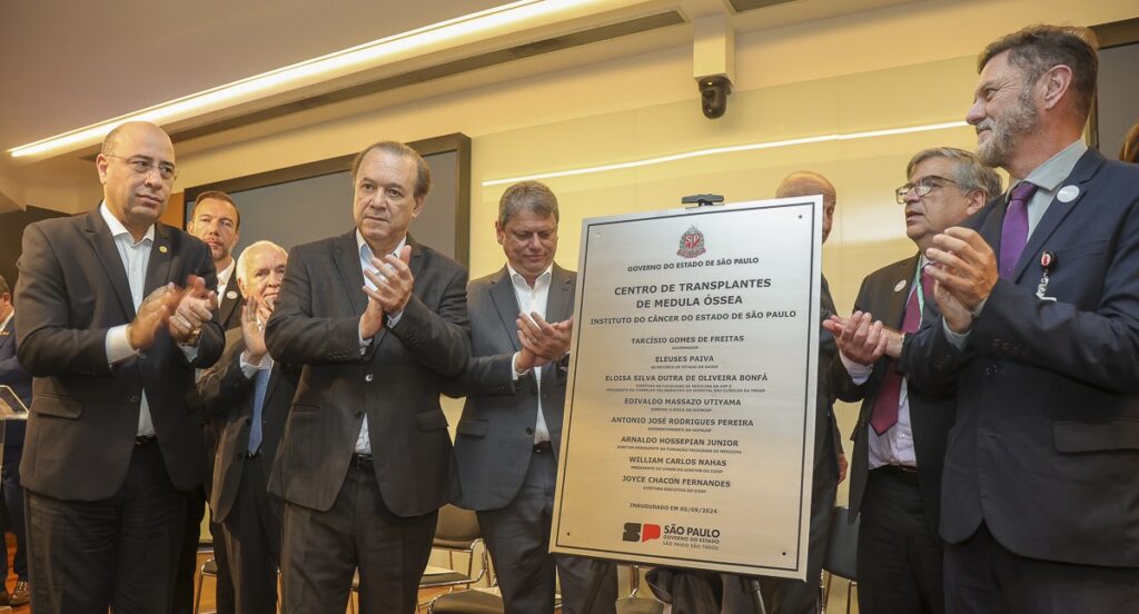 Homens em pé, ao lado da placa de metal, aplaudem o descerramento da placa de inauguração do Centro de Transplantes de Medula Óssea.