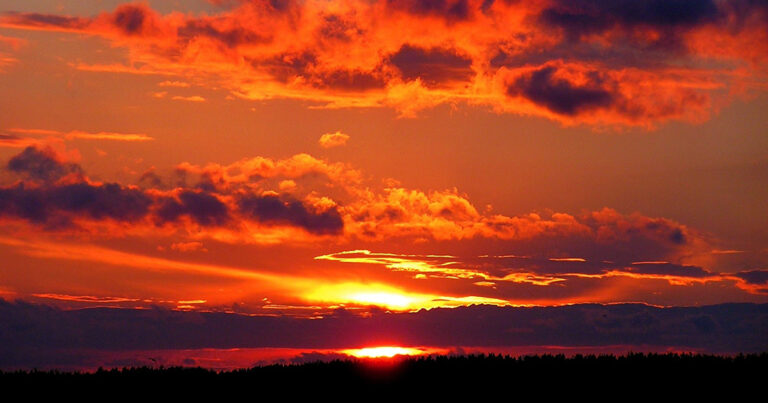 A imagem mostra um pôr do sol emitindo uma luz que ilumina as nuvens ao redor em tons de laranja, vermelho e roxo