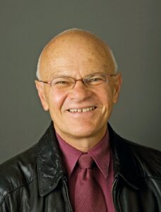 O professor e escritor Leo Spitzer, homem branco, sorrindo, com óculos de grau, camisa e gravata na cor magenta/vinho e jaqueta de couro preta