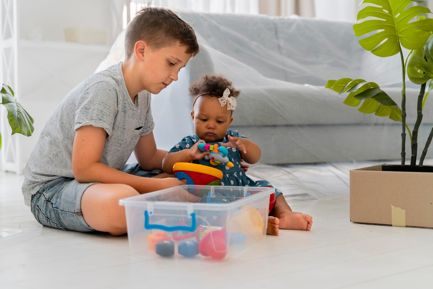 Uma criança branca brincando com uma bebê negra com briquedos de plástico no chão. À direita há uma planta