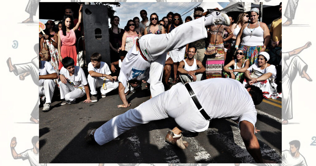 Na imagem dois homens vestidos de branco aparecem em movimento de capoeira, um agachado e de costas com a perna esquerda esticada e outro de cabeça para baixo com as duas mãos e a perna direita no chão e a perna esquerda para cima. Ao fundo aparecem vários expectadores. 