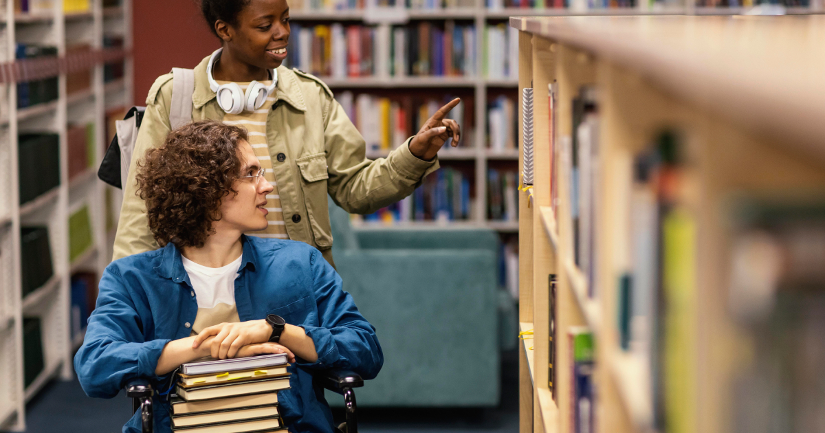 homem jovem branco na cadeira de rodas com uma mulher negra jovem mostrando os livros em uma biblioteca.