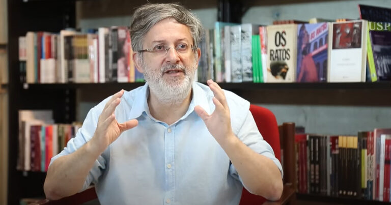 Homem de meia idade, com cabelo e barba branca e com estante de livros ao fundo