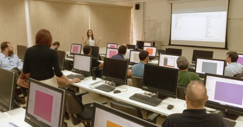 Sala de aula com pessoas e computadores nas mesas e uma pessoa dando aula com uma tela de projeção na frente da sala