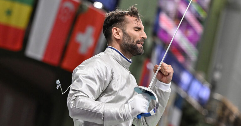 Medalhista olímpico branco de cabelo liso, segurando espada de esgrima e comemorando