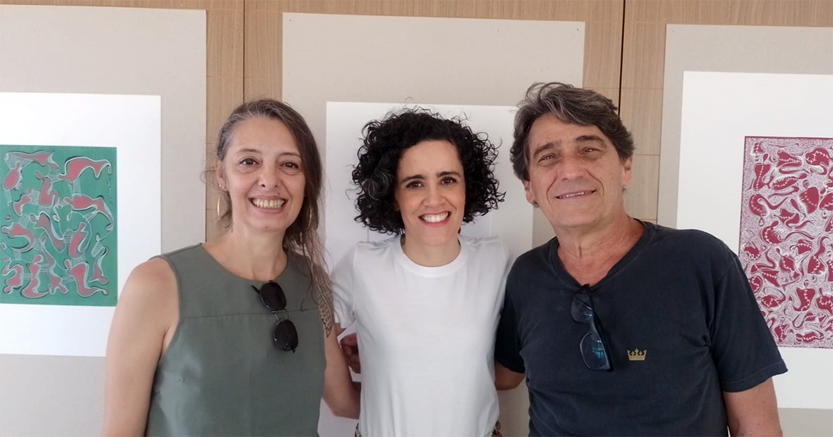 Dois artistas abraçados com uma realizadora do evento, ao fundo pinturas