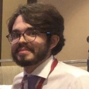 foto de um jovem branco, com cabelos e barba escuras. Ele usa óculos, camisa social branca e está de perfil esboçando um sorriso.