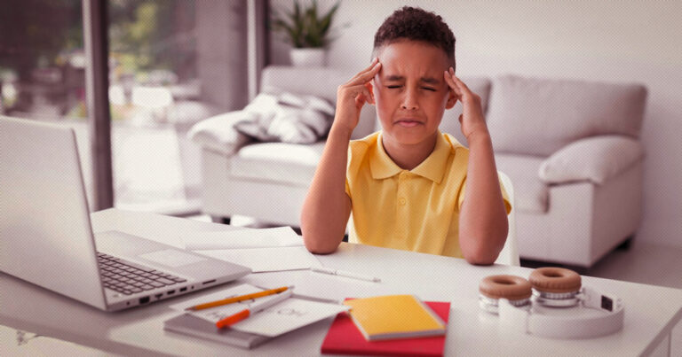 foto mostra um menino de camisa polo amarela com as duas mãos nas têmporas. Ele está sentado em frente a uma em uma bancada, onde estão um laptop aberto, blocos de anotações, canetas e lápis.