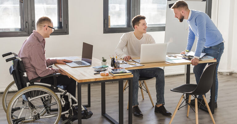 Três homens brancos conversam em um escritório. Um deles está em pé, à direita, enquanto os outros dois estão à mesa. trabalhando em seus computadores. O homem à esquerda usa cadeira de rodas.