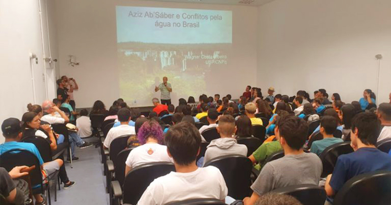 Várias pessoas sentadas em cadeiras assistindo uma palestra com um professor e uma projeção na parede atrás dele