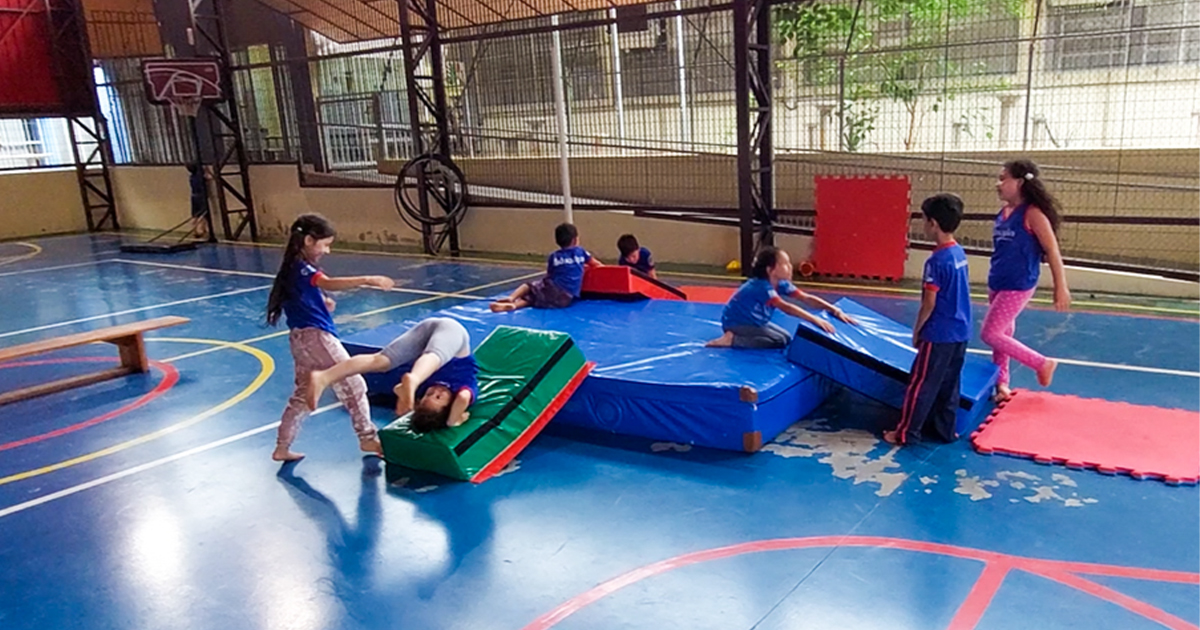 crianças fazem ginástica na quadra de uma escola, brincando em rampas e tapetes coloridos