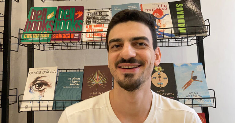 homem branco de cabelo liso, sorrindo olhando para foto, atrás dele uma estante de livros