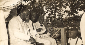 Imagem: Grupo de homen negros, todos vestindo trajes brancos, e tocando instrumentos de percussão