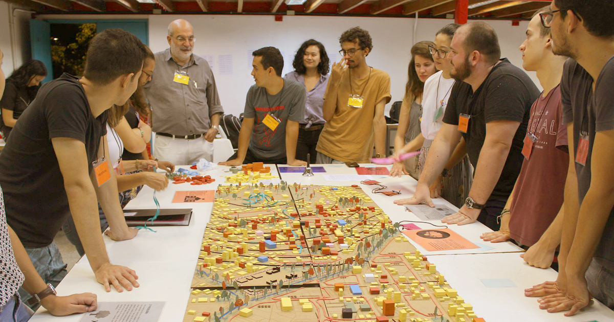 Pessoas em volta de uma mesa com um jogo que tem bloquinhos que parecem prédios e casas