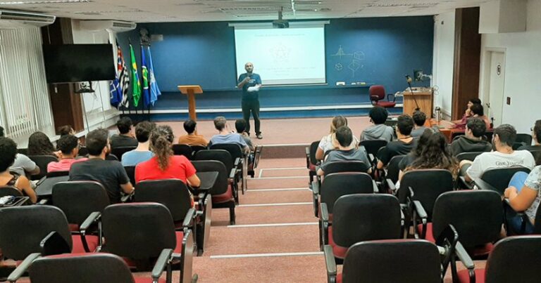 Sala de aula com alunos sentados e professor falando e ao fundo uma lousa