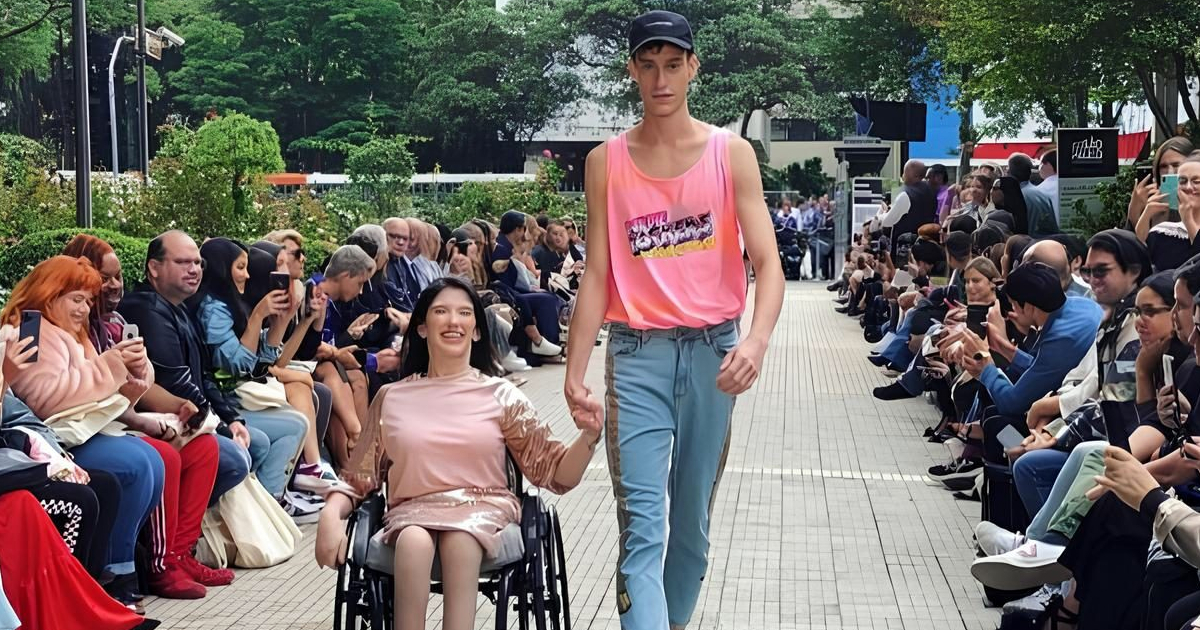 Imagem: Homem branco desfilando em passarela de mãos dadas com uma jovem e sorridente cadeirante