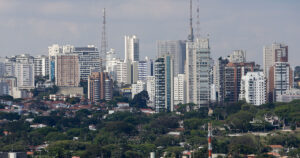 Vista de uma grande cidade mostrando uma área com árvores na parte de baixo e prédios e torres na parte cima. O céu está nublado