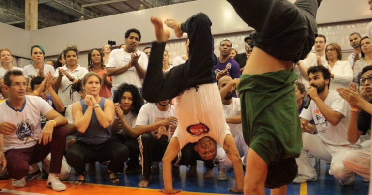 duas pessoas jogando capoeira diante de um grupo que bate palmas