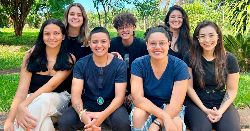 Grupo de sete pessoas quatro sentadas na frente e 3 atrás com as mãos nos joelhos, duas pessoas na frente e ao centro com camisetas azuis e as demais com camisetas pretas 