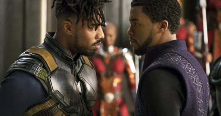 A imagem mostra uma cena do filme "Pantera Negra" (2018), da Marvel Studios. Nela, os personagens Erik Killmonger, interpretado por Michael B. Jordan, e T'Challa, interpretado por Chadwick Boseman, estão se encarando de perto em uma pose tensa e confrontativa. Killmonger está usando uma armadura com detalhes dourados e T'Challa está vestindo um traje roxo com padrões geométricos. Ao fundo, há uma figura desfocada em trajes vermelhos