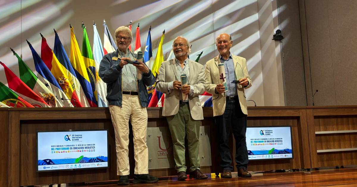 Três educomunicadores sorridentes estão em pé sobre um palco, com seus prêmios nas mãos. Os três são brancos e idosos.