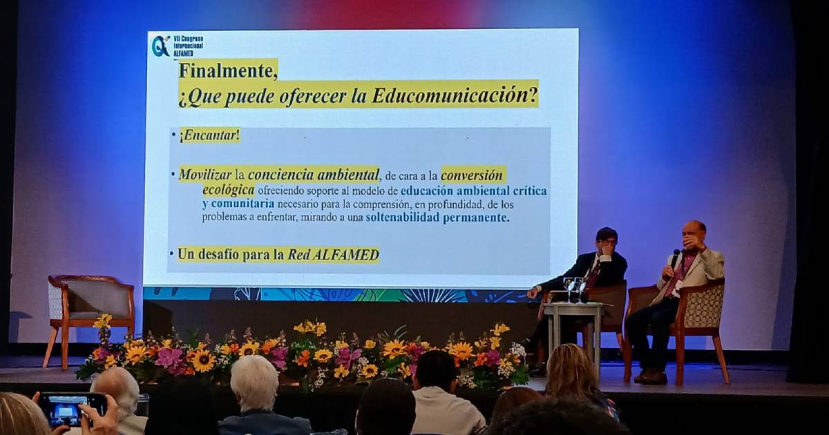 O palestrante e o mediador da palestra estão sentados em poltronas colocadas na ponta direita do telão da apresentação