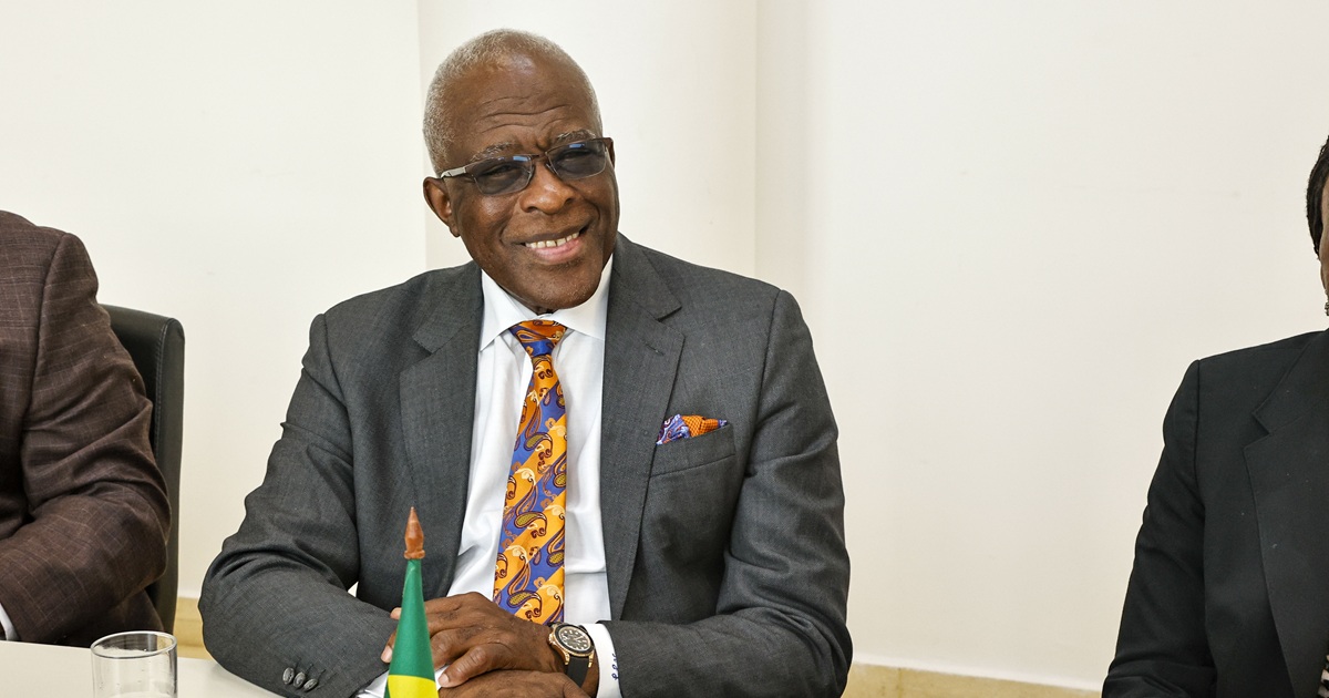 Homem negro, com paletó cinza, gravata colorida, lenço igual no bolso do paletó, óculos pretos, sorridente, sentado à uma mesa, com uma pequena bandeira do Brasil à frente