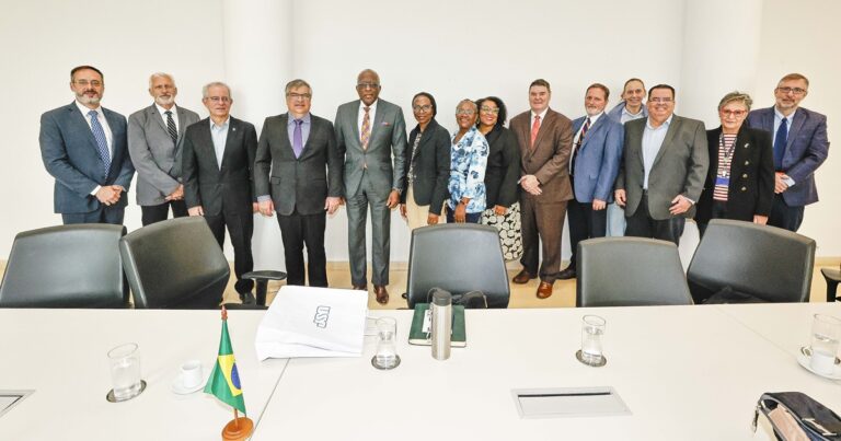 Grupo com várias pessoas posando para foto em uma sala grande. À frente, uma mesa com copos, cadernos, uma pequena bandeira do Brasil