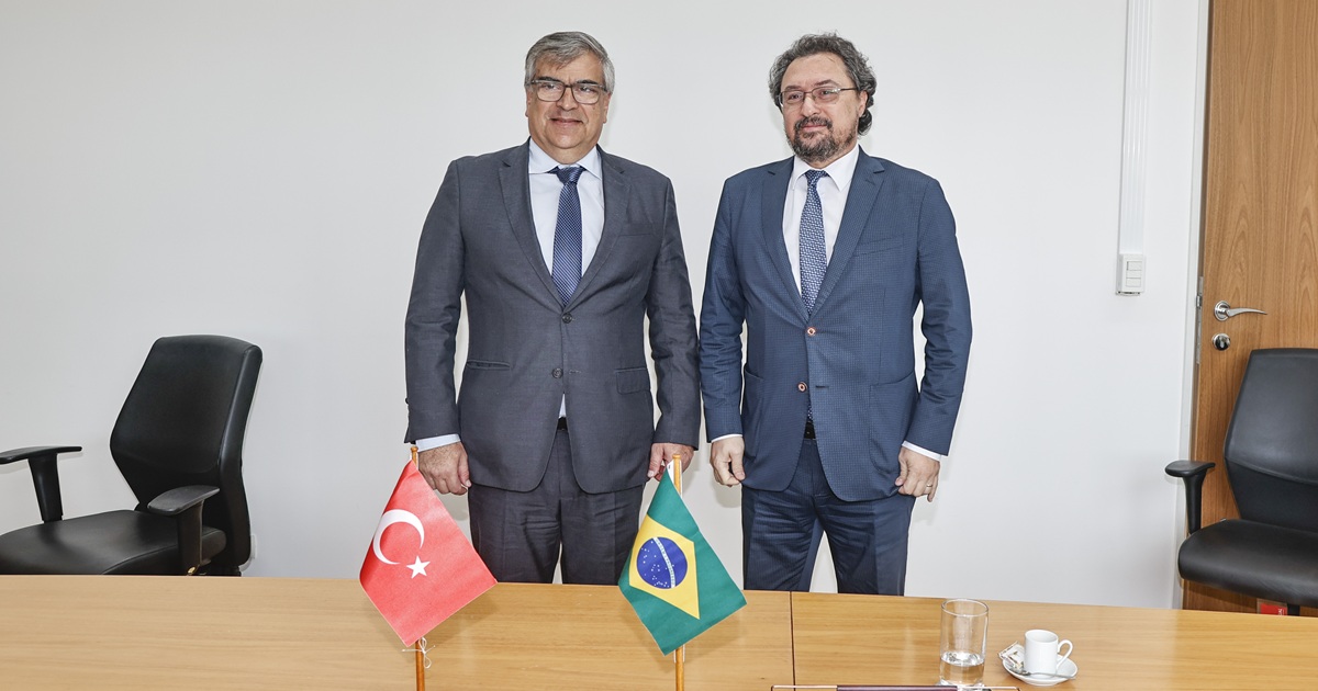 Dois homens, de terno e gravata, posam de pé. À frente, uma mesa com pequenas bandeiras da Turquia e do Brasil