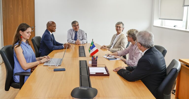 Mesa de madeira, com pequenas bandeiras da França e do Brasil. Seis pessoas estão sentadas ao redor da mesa