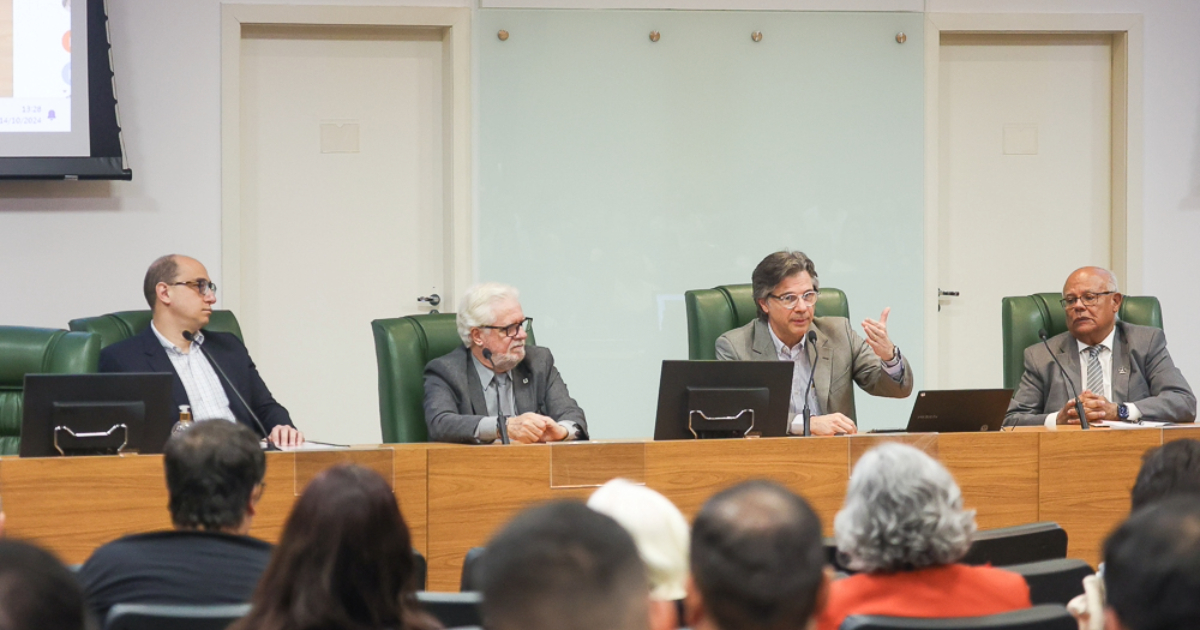 Quatro homens sentados à mesa principal do evento, falando para a plateia