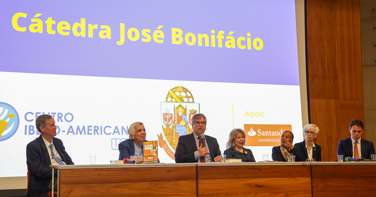 Grupo de sete pessoas, sentado, à uma mesa em cima do palco