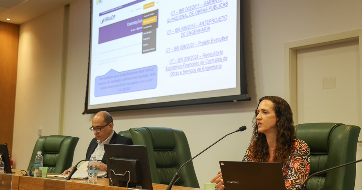 Um homem de óculos e uma mulher de cabelos longos na mesa do evento. Atrás deles, um telão exibe informações que estão sendo apresentadas no treinamento.