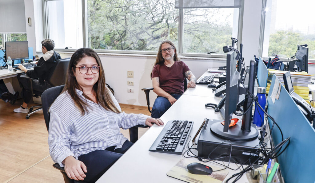 Mulher branca, de óculos, cabelo longo e camisa listrada, ao lado de um homem branco, de óculos, cabelo longo e camiseta vinho. Ambos sentados na frente de seus respectivos computadores.