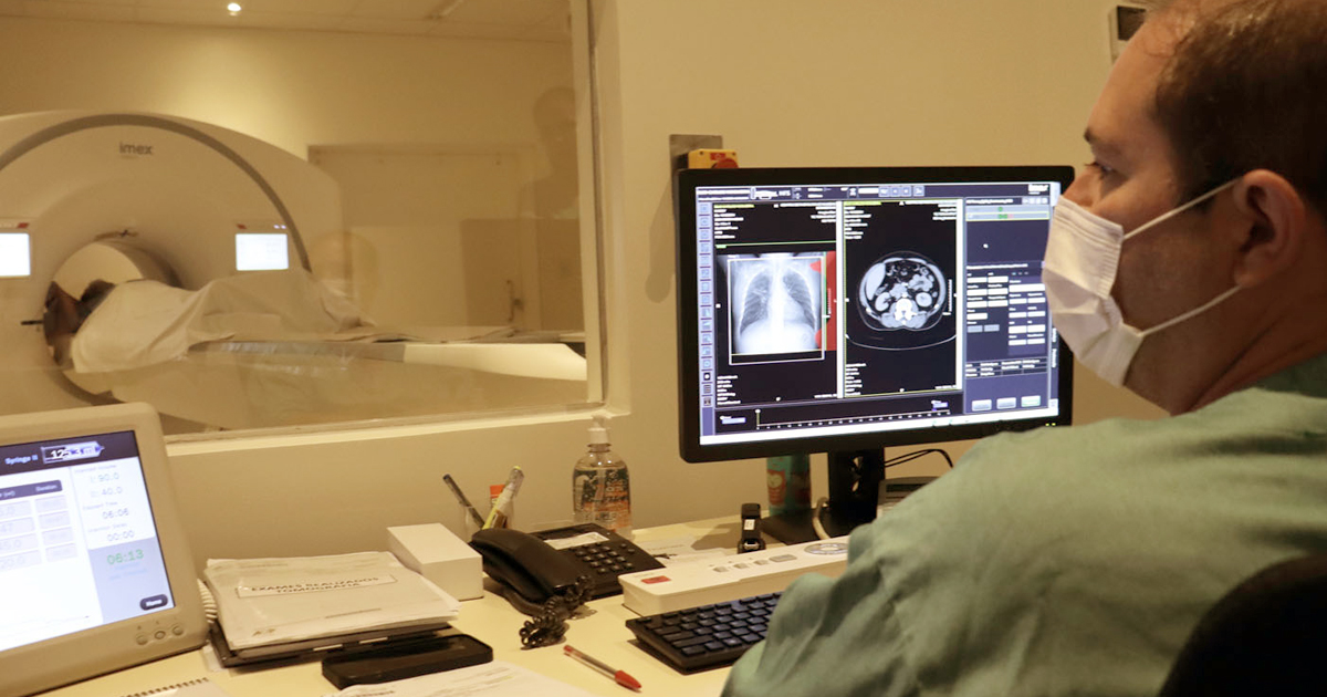 Técnico realizando exame de tomografia com a tela do computador à frente mostrando imagens e o tomógrafo ao fundo