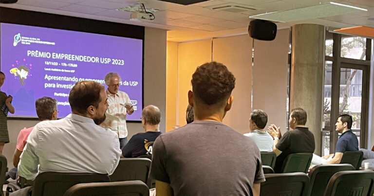 Sala de evento, com várias pessoas sentadas e um homem dando palestra, tendo ao fundo um telão