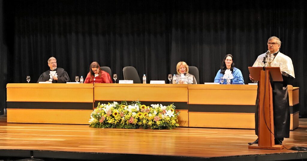 Na imagem, mesa clara com 3 mulheres e um homem sentados, e um homem em pé num púlpito com roupas pretas e capa branca