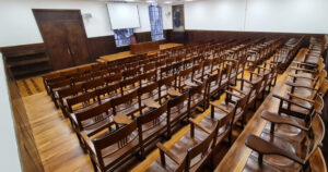 Sala de aula, com cadeiras de madeira com braços, ao fundo portas e janelas