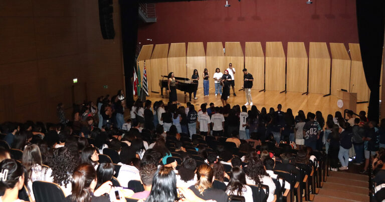 Auditório cheio de alunos sentados e uma fileira na beirada do palco, onde tem mais 6 alunos eum num piano de cauda. todos fotografados do fundo e a luz incidindo somente no palco