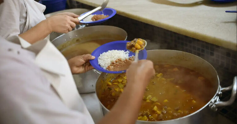 Foco nas mãos de duas pessoas servindo pratos de arroz, feijão e picadinho de carne a partir de grandes panelas num balcão