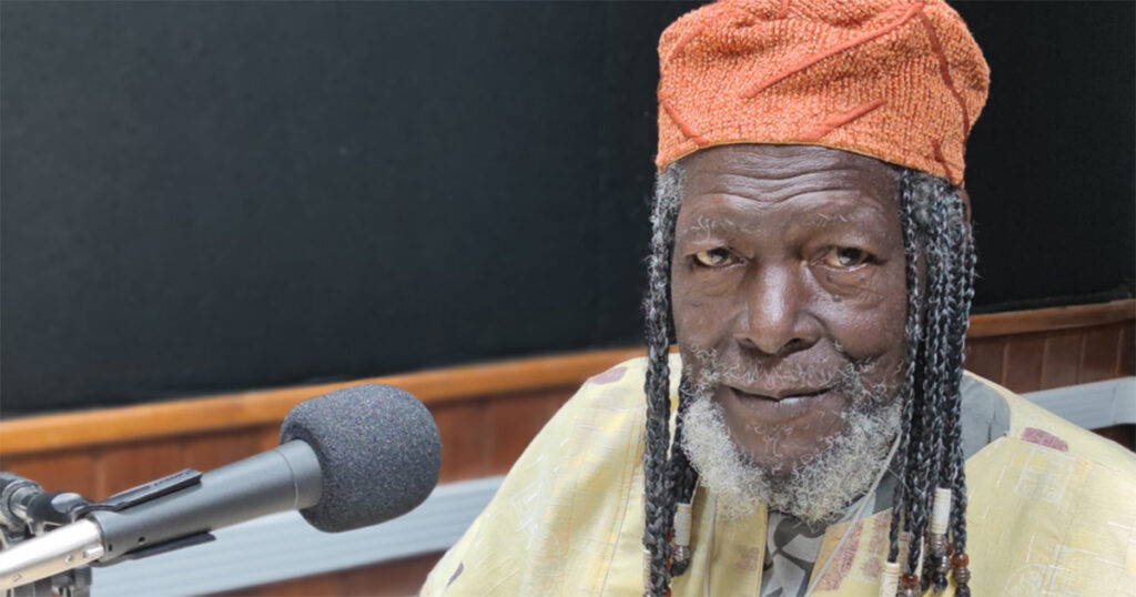 Artista plástico Macalé, homem negro com barba e cabelo longo com dreds, usando gorro laranja posando para foto em frente a um microfone