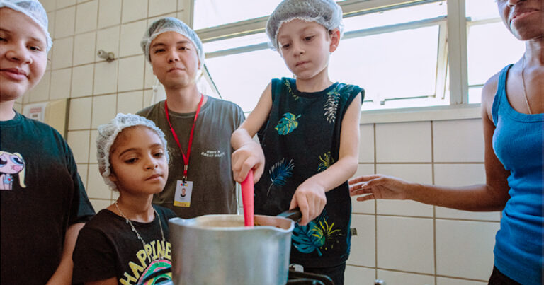 Crianças numa cozinha de escola sendo uma delas mexendo a comida numa panela